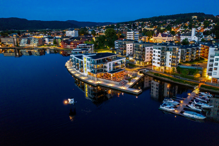 Øya Spiseri Porsgrunn – restaurant ved vannkanten sett fra luften, med opplyst uteservering og utsikt over Porsgrunnselva.