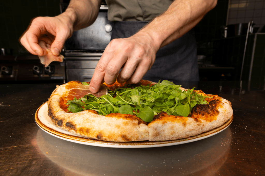 Øya Spiseri Porsgrunn – kokk som tilbereder italiensk-inspirert pizza med ruccola og spekeskinke.