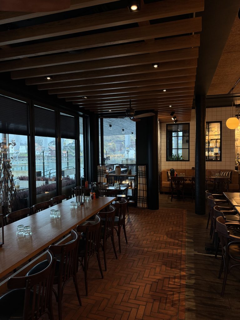 Øya Spiseri Porsgrunn – moderne restaurantinteriør med langbord, varme trematerialer og utsikt over Porsgrunnselva gjennom store vinduer.