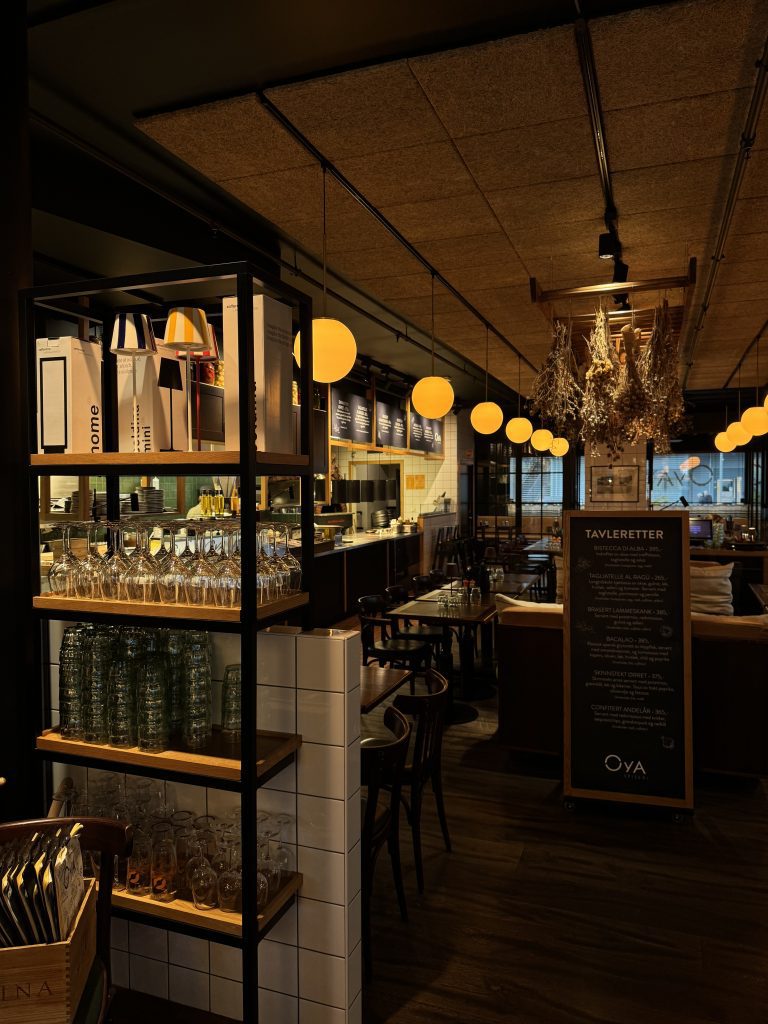 Øya Spiseri Porsgrunn – varm og moderne restaurantatmosfære med åpne hyller, vinglass, gule pendellamper og innsyn til kjøkken og spiseplasser.